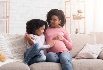 Curious little daughter touching her pregnant mother tummy, sitting on sofa at home, empty space