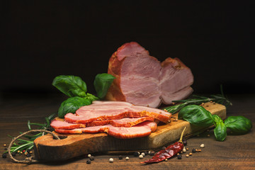 Sliced smoked pork meat with spices lies on an ancient cutting Board on a wooden rustic table against a dark background. Nearby are rosemary and Basil. Focus concept