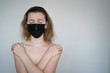 a naked girl in a black medical mask crossed her arms over her chest against a white background. The concept of defenselessness from the coronavirus. space for text
