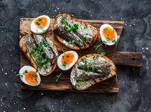 Delicious Appetizer, Tapas - Soft Boiled Eggs And Canned Sardines Sandwiches On A Cutting Board On A Dark Background, Top View