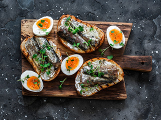 Wall Mural - Delicious appetizer, tapas - soft boiled eggs and canned sardines sandwiches on a cutting board on a dark background, top view