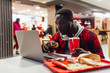 © Тарас Нагирняк - Photo of serious freelancer, african man is sitting at modern cafe, use laptop while eating fast food and drinking cola.