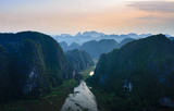 Fototapeta Góry - Aerial view at sunset of Ninh Binh region, Tam Coc valley tourist attraction, UNESCO World Heritage Site, scenic river crawling through karst mountain ranges in Vietnam, travel destination.