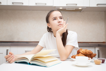 A young woman at the table plans a day with a pen and notebook, has breakfast, drinks coffee.