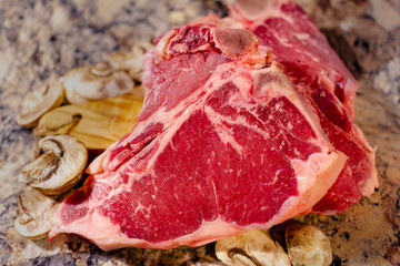 A T bone or Porterhouse raw steak on cutting board with mushrooms.  Blurred foreground.  Blurred background.  Neutral marbled surface.