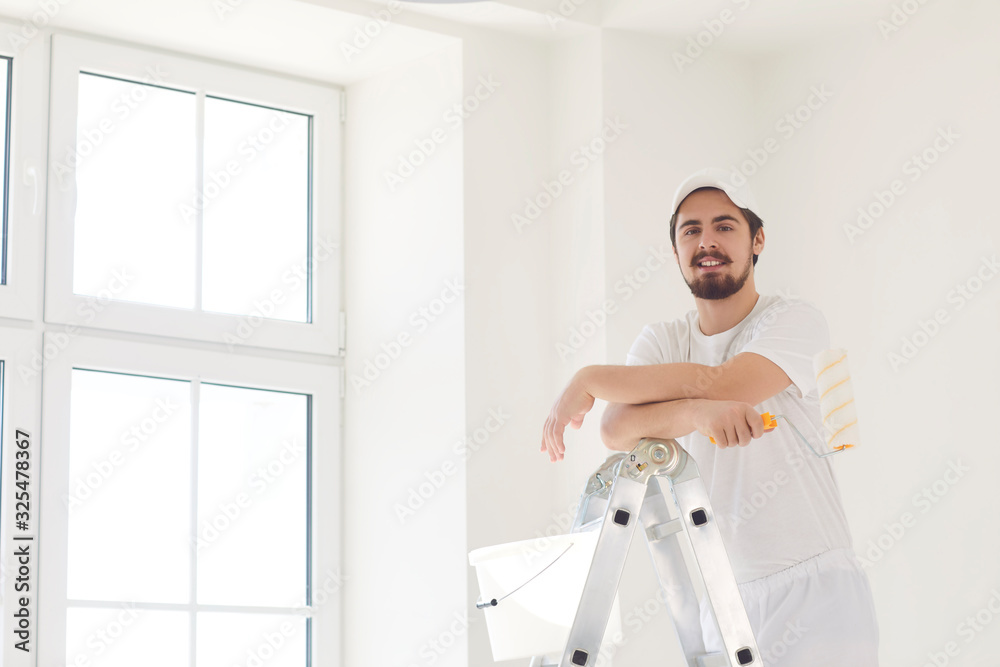 A male painter in a white uniform with a roller works in his hand in a white room