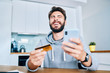 © baranq - Portrait of cheerful young man laughing while paying online from home with credit card and smartphone