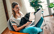 © Valerii Apetroaiei - An attractive young woman in glasses is working on a laptop while sitting cross-legged in a comfortable chair at home with a funny assistant cat on her legs.