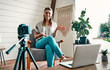 © Valerii Apetroaiei - Attractive young woman blogger dressed in jeans and a blouse is recording on the camera while sitting on a comfortable chair at home.