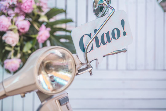 Word Ciao written on a bubble text board stuck behind the handlebar of an italian moto scooter with visible headlamp. White boards in the background and pink flowers.