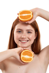 Fruits for skin care. Portrait of young and happy red-haired girl with freckles on face holding two pieces of orange and smiling at camera while standing against white background