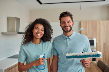 Wall Mural - Keeping your home super clean. Happy male and female professional cleaners in uniform holding cleaning supplies, looking at camera and smiling while standing in the kitchen. Cleaning service