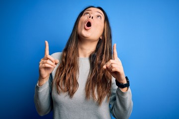 Young beautiful brunette woman wearing casual sweater standing over blue background amazed and surprised looking up and pointing with fingers and raised arms.