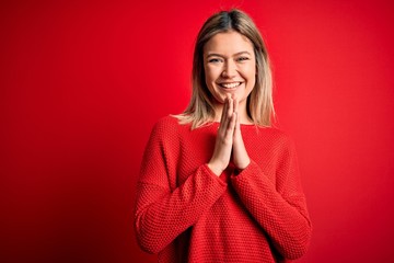 Poster - Young beautiful blonde woman wearing casual sweater over red isolated background praying with hands together asking for forgiveness smiling confident.