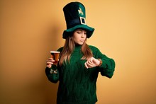 Young Beautiful Woman Wearing Green Hat Drinking Glass Of Beer On Saint Patricks Day Checking The Time On Wrist Watch, Relaxed And Confident