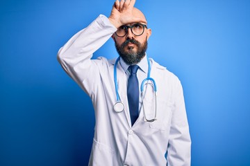 Sticker - Handsome bald doctor man with beard wearing glasses and stethoscope over blue background making fun of people with fingers on forehead doing loser gesture mocking and insulting.