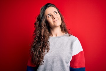 Young beautiful woman with curly hair wearing casual sweatshirt over isolated red background smiling looking to the side and staring away thinking.