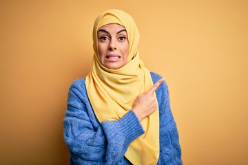 Poster - Young beautiful brunette muslim woman wearing arab hijab over isolated yellow background Pointing aside worried and nervous with forefinger, concerned and surprised expression