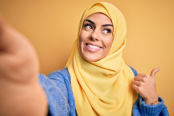 Poster - Young beautiful brunette arab woman wearing islamic hijab make selfie by camera pointing and showing with thumb up to the side with happy face smiling