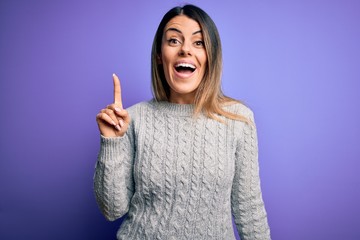 Canvas Print - Young beautiful woman wearing casual sweater standing over isolated purple background pointing finger up with successful idea. Exited and happy. Number one.