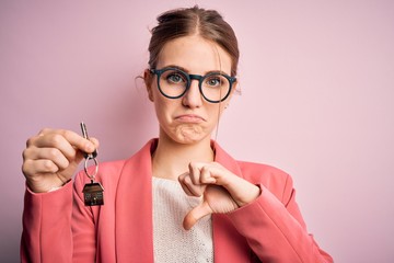 Young beautiful redhead house agent woman holding home key over pink bakcground with angry face, negative sign showing dislike with thumbs down, rejection concept