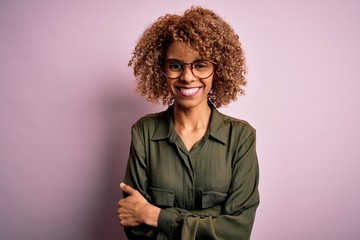 Young beautiful african american woman wearing casual shirt and glasses over pink background happy face smiling with crossed arms looking at the camera. Positive person.