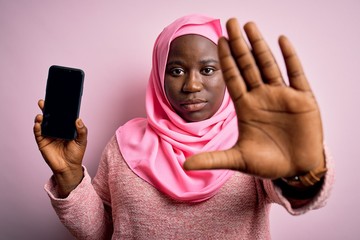 Poster - African american plus size woman wearing muslim hijab holding smartphone showing screen with open hand doing stop sign with serious and confident expression, defense gesture