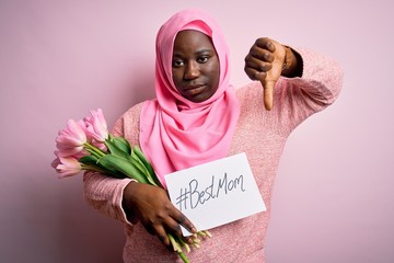 Wall Mural - Plus size african american woman wearing hijab holding message and tulips on mothers day with angry face, negative sign showing dislike with thumbs down, rejection concept