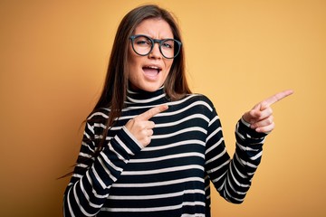 Beautiful woman with blue eyes wearing striped sweater and glasses over yellow background smiling and looking at the camera pointing with two hands and fingers to the side.