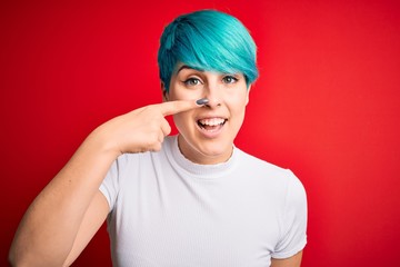 Young beautiful woman with blue fashion hair wearing casual t-shirt over red background Pointing with hand finger to face and nose, smiling cheerful. Beauty concept