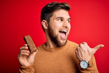 Sticker - Young blond man with beard and blue eyes eating healthy chocolate protein bar pointing and showing with thumb up to the side with happy face smiling