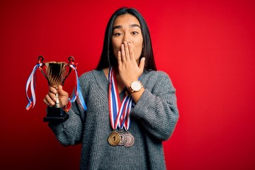 Sticker - Young beautiful champion asian woman holding trophy wearing medals over red background cover mouth with hand shocked with shame for mistake, expression of fear, scared in silence, secret concept