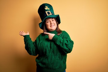 Wall Mural - Young beautiful plus size woman wearing green hat with clover celebrating saint patricks day amazed and smiling to the camera while presenting with hand and pointing with finger.