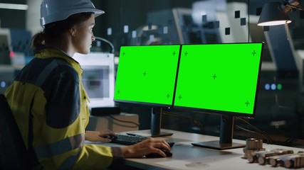 Poster - Female Engineer Wearing Safety Vest and Hardhat Works on a Computer, Two Monitor Screens Show Chroma Key / Green Screen Displays. Modern High-Tech Industry 4.0 Electronics Production Factory