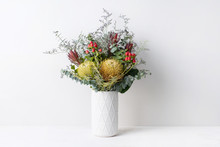 Elegant Flower Arrangement Of A Dried Banksia Surrounded By Misty Blue, Red Leucadendrons And Australian Eucalyptus, In A White Vase On A Table. Could Be A Gift On Display Or Decoration.