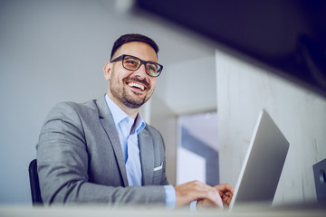 Cheerful caucasian classy unshaven businessman in suit and with eyeglasses using laptop while sitting in his office.