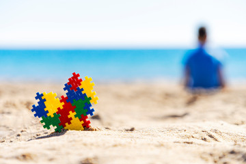 Colorful autism awareness heart on the sand and back view to silhouette of the child sitting on the beach and looking to the sea ocean