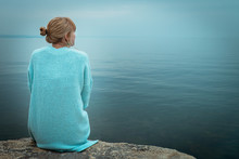 Beautiful Single Solo Young Traveler Blond Woman With Tranquit Jacket Sitting Alone On Stone Looking In To Water In The Blue Ocean Foggy Mist Lake In Cold Winter With Sad Dark Depression  Emotional