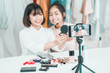 © sutthinon602 - Two young girls doing video recording by smartphone camera. Asian woman make up artist applying powder on female blogger face with cosmetic products table in living room. Beauty ladies live stream