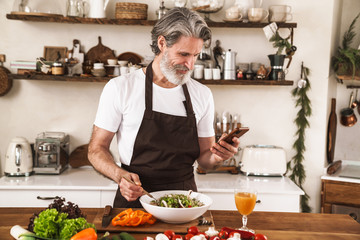 Image of pleased gray-haired man using cellphone and preparing salad