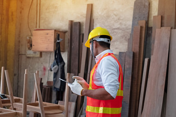 Handsome man wearing safety vest and yellow helmet,holding tablet in hand,for checking wood work at factory,blurry light around