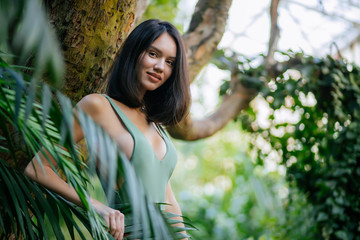 Wall Mural - good-looking slim caucasian female in green fashionable swimsuit, posing around green exotic plants in jungle, in tropical rainforest. fashion models concept