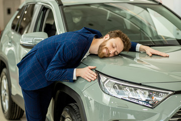 portrait of positive smiling happy man buyer in dealership, stand happily posing next to new car, wearing formal suit. funny bearded man hug new auto