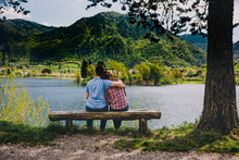 Young Couple Sitting On Bench Under A Tree At Lake In Italy.