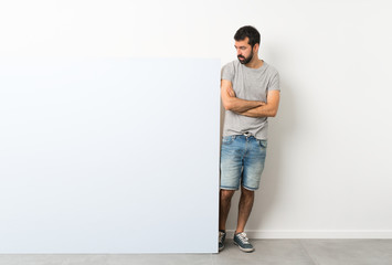 Young handsome man with beard holding a big blue empty placard with confuse face expression