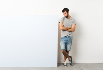 Wall Mural - Young handsome man with beard holding a big blue empty placard feeling upset