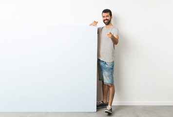 Wall Mural - Young handsome man with beard holding a big blue empty placard points finger at you while smiling