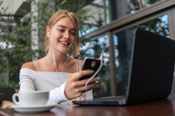 Modern business woman is reading incoming sms message on smartphone or watching broadcasting online on modern mobile phone while sitting in caffe with a laptop. Female student sitting in coffee.