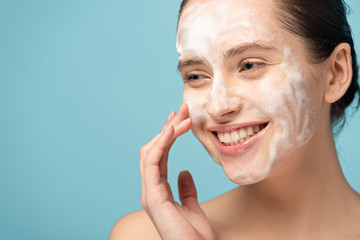 Wall Mural - beautiful smiling woman applying cleansing foam on face, isolated on blue