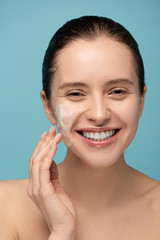 Wall Mural - smiling girl applying cleansing foam on face, isolated on blue